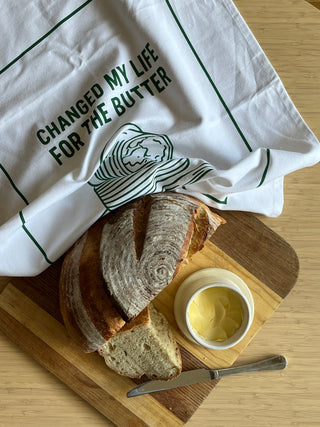 Loaf of bread with a slice cut, butter dish, and knife on a wooden cutting board with a tea towel.